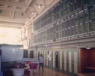 An interior space with a high ceiling featuring a large electronic board displaying various financial data and numbers. There is modern seating with red and white chairs, and a person is walking through the room.