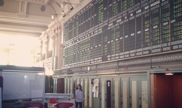An interior space with a high ceiling featuring a large electronic board displaying various financial data and numbers. There is modern seating with red and white chairs, and a person is walking through the room.