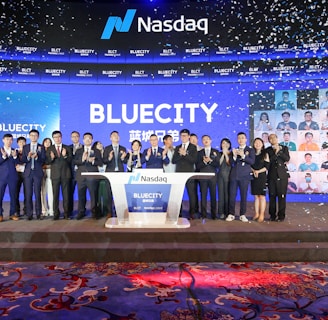 A group of people dressed in formal attire stands on a stage with a Nasdaq podium in the center. The backdrop displays the word 'BLUECITY' along with various smaller images of individuals. Confetti is falling, creating a celebratory atmosphere.