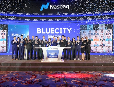 A group of people dressed in formal attire stands on a stage with a Nasdaq podium in the center. The backdrop displays the word 'BLUECITY' along with various smaller images of individuals. Confetti is falling, creating a celebratory atmosphere.