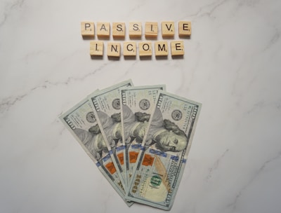 A marble surface displays five 100-dollar bills arranged in a fan shape. Above the bills, the words PASSIVE INCOME are spelled using wooden Scrabble tiles.