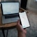 A person holds a smartphone displaying a stock market trading app with candlestick charts. A laptop is on a wooden table with a similar financial chart, indicating active trading or analysis. The setting suggests a home office or casual workspace.