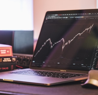 A laptop displaying a stock market graph with a rising trend is placed on a desk. Next to the laptop are several books stacked on each other, with titles related to investing. A mouse is positioned to the right of the laptop.