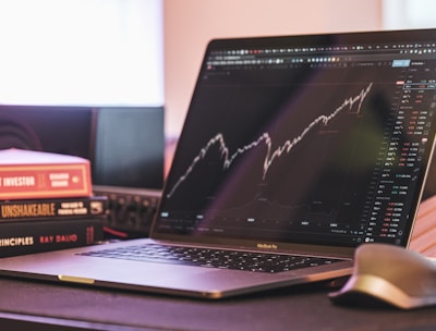A laptop displaying a stock market graph with a rising trend is placed on a desk. Next to the laptop are several books stacked on each other, with titles related to investing. A mouse is positioned to the right of the laptop.