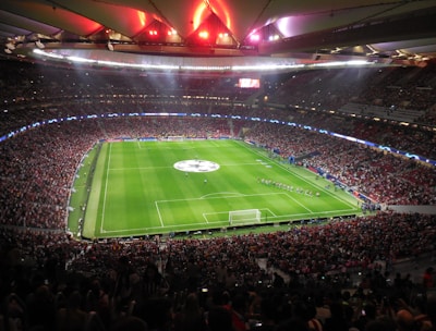 A large, packed stadium with bright lights illuminates a green soccer field. The scene is vibrant with a massive audience surrounding the pitch, visibly excited and engaged. The center of the field displays a prominent circular logo, suggesting a major sporting event. Players and staff can be seen on the field, likely preparing for a match.