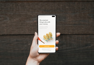 A hand holding a smartphone in front of a wooden background. The phone screen displays a financial investment application with the text 'The smartest thing to do with your money'. Below it, there are illustrations of stacked coins and a call-to-action button labeled 'Start investing'.