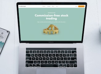 A laptop with a financial website open is centered on a white table. The screen displays a mint green background with text related to commission-free stock trading, accompanied by a gold object resembling a bridge. To the left, a smartphone displays a financial app with a graph and monetary figure. A cup of coffee sits on the right side, adding a casual work environment atmosphere.