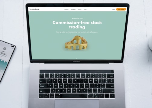 A laptop with a financial website open is centered on a white table. The screen displays a mint green background with text related to commission-free stock trading, accompanied by a gold object resembling a bridge. To the left, a smartphone displays a financial app with a graph and monetary figure. A cup of coffee sits on the right side, adding a casual work environment atmosphere.