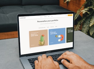 A person is typing on a MacBook Pro, viewing a website titled 'Personalize your portfolio.' The webpage displays three sections related to investing, each with different images and text. The laptop is on a grey table with a dark sofa and indoor plants in the background.