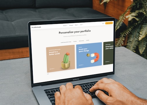 A person is typing on a MacBook Pro, viewing a website titled 'Personalize your portfolio.' The webpage displays three sections related to investing, each with different images and text. The laptop is on a grey table with a dark sofa and indoor plants in the background.