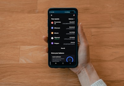 A hand holds a smartphone displaying a cryptocurrency portfolio with various assets including Decentraland, Ethereum, Bitcoin, Algorand, and Polygon. The balance for each asset is shown alongside icons representing each cryptocurrency.