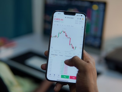 A person holds a smartphone displaying a financial or stock trading app, with detailed graphs and numerical data visible on the screen. In the background, there is a blurred computer monitor and office setting, suggesting a work environment.