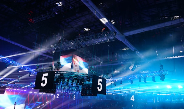 A large indoor arena is equipped with advanced lighting and audio systems. Suspended above the space are numerous digital displays showcasing numbers. The environment is filled with blue illumination and spotlights creating a vibrant atmosphere.