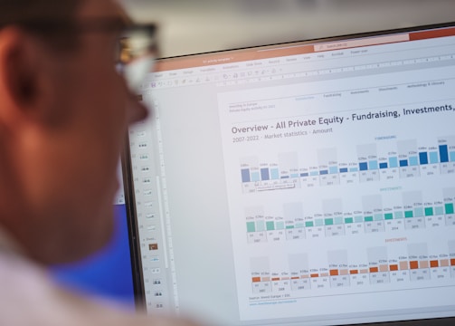 A person is looking at a computer screen displaying a document titled 'Overview - All Private Equity - Fundraising, Investments, & D'. The document contains statistical charts and graphs related to private equity market statistics from 2007 to 2022. Various tabs and functions like 'File', 'Home', 'Insert', and 'Design' are visible in the interface.