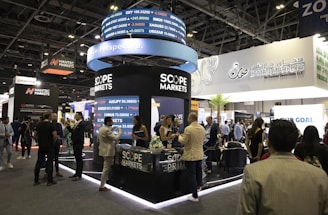 A bustling exhibition hall filled with people attending a financial conference or trade event. Prominent displays and booths from companies like Scope Markets, Daman Markets, and Hantec Markets are visible. Attendees are engaged in conversations, networking, and exploring the offerings and information provided by the exhibitors.