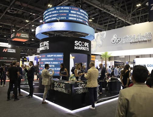 A bustling exhibition hall filled with people attending a financial conference or trade event. Prominent displays and booths from companies like Scope Markets, Daman Markets, and Hantec Markets are visible. Attendees are engaged in conversations, networking, and exploring the offerings and information provided by the exhibitors.