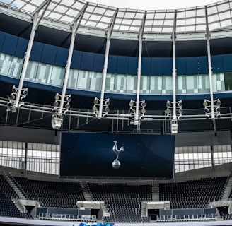 A modern stadium interior featuring a large digital screen with a logo prominently displayed. The seating areas are empty, with dark-colored seats and some structural elements like beams and glass walls visible. A combination of dark blue and neutral tones are prominent.