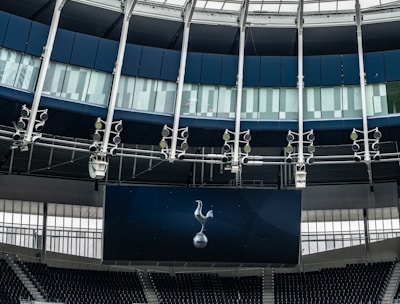 A modern stadium interior featuring a large digital screen with a logo prominently displayed. The seating areas are empty, with dark-colored seats and some structural elements like beams and glass walls visible. A combination of dark blue and neutral tones are prominent.